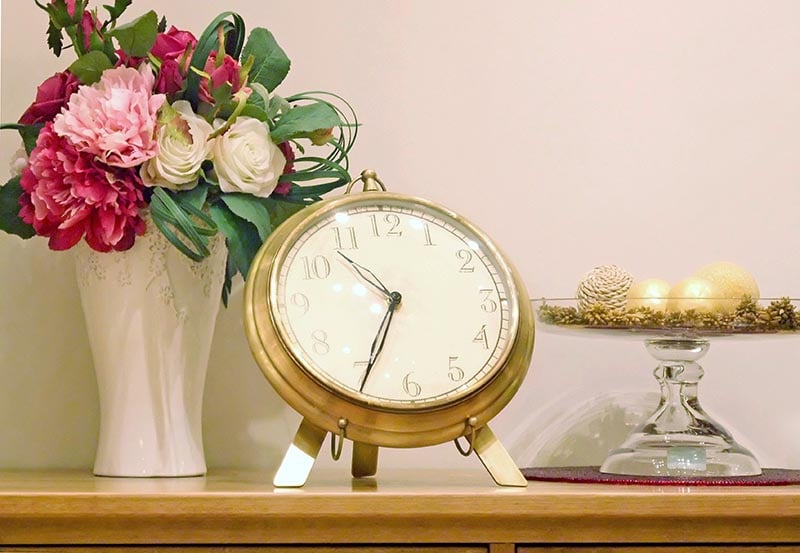 A vintage brass tabletop clock displayed alongside a vase of pink and white flowers and decorative ornaments on a wooden shelf.