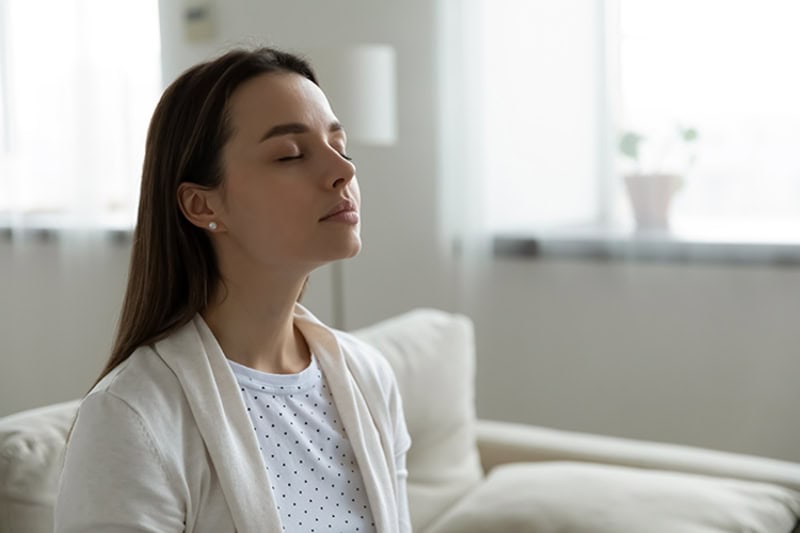 A calm woman sitting on a couch with eyes closed, enjoying fresh air and indoor comfort.