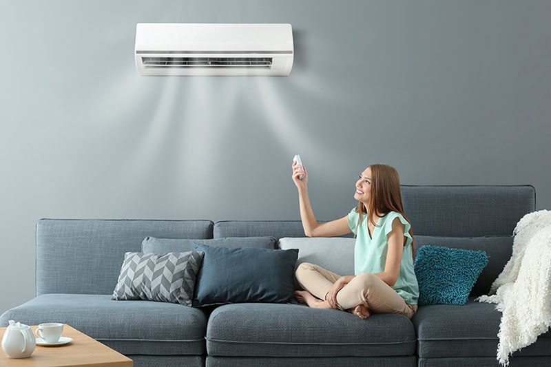 Hot and Cold Spots? Get Even Heating and Cooling. Photo of a young woman switching on the AC with a remote while sitting on a sofa in her home.