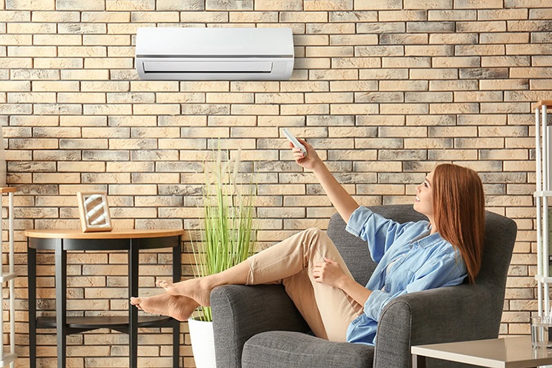 How to Cool the Upstairs of a Two-Story Home. Photo of a young woman sitting in an arm chair switching the AC on with a remote.