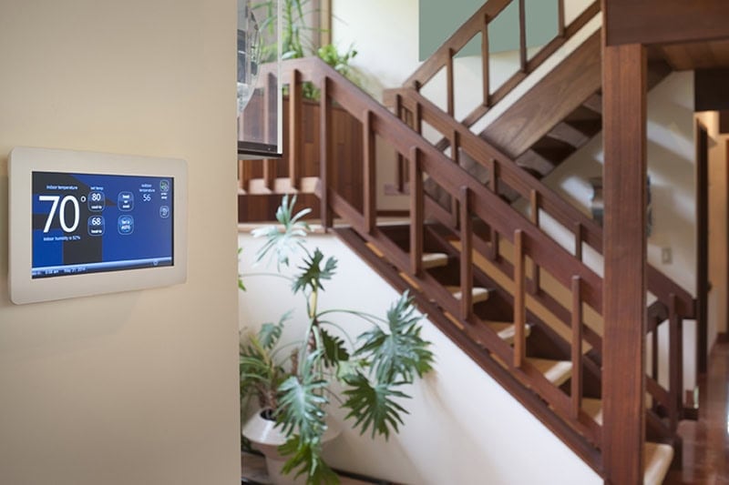 How a Smart Thermostat Can Save You Money During a Heat Wave. Photo of a digital thermostat mounted on a wall with a staircase and plant in the background.